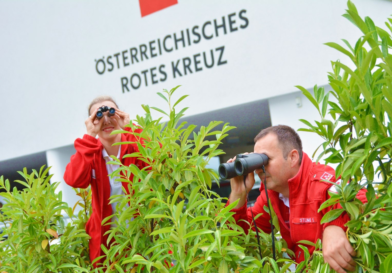 © Rotes Kreuz Kirchdorf Ein Rotkreuz-Mitarbeiter und eine Rotkreuz-Mitarbeiterin verstecken sich humorvoll und schauen durch ein Fernglas. Sie suchen etwas.