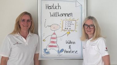 Zwei Frauen stehen vor einem Plakat, auf dem Herzlich Willkommen steht. Die Frauen sind Trainerinnen eines Workshops für psychische Erste Hilfe.