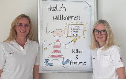 Zwei Frauen stehen vor einem Plakat, auf dem Herzlich Willkommen steht. Die Frauen sind Trainerinnen eines Workshops für psychische Erste Hilfe.