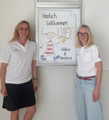 Zwei Frauen stehen vor einem Plakat, auf dem Herzlich Willkommen steht. Die Frauen sind Trainerinnen eines Workshops für psychische Erste Hilfe.