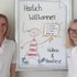 Zwei Frauen stehen vor einem Plakat, auf dem Herzlich Willkommen steht. Die Frauen sind Trainerinnen eines Workshops für psychische Erste Hilfe.