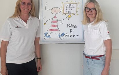 Zwei Frauen stehen vor einem Plakat, auf dem Herzlich Willkommen steht. Die Frauen sind Trainerinnen eines Workshops für psychische Erste Hilfe.