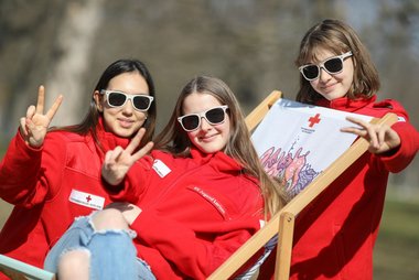 Drei Mädchen in Rotkreuz-Jacke und mit Jugendrotkreuz-Sonnenbrille lachen in die Kamera.