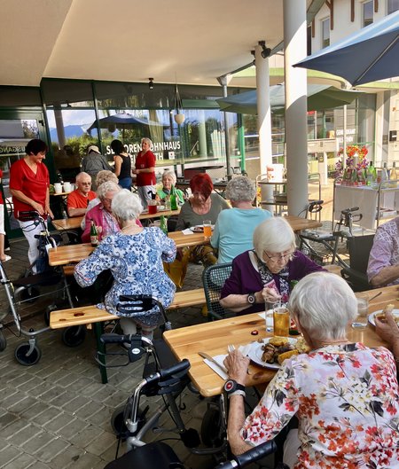 Bewohnerinnen und Bewohner sitzen an Tischen und genießen Essen und Getränke während des Grillfestes