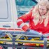 eine junge blonde Frau steht in Rotkreuz-Uniform vor einem Rettungswagen und putzt die Trage des Rettungswagens