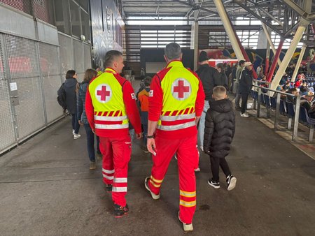 Eine Gruppe von Kindern geht mit dem Rücken zur Kamera durch das Stadion, hinter ihnen und damit näher zur Kamera zwei erwachsene Männer in Rotkreuz-Uniform mit gelber Weste.