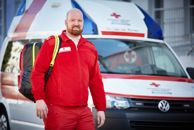 Ein männlicher Rettungssanitäter, steht vor einem Rettungsauto mit einem Rucksack über die Schulter.