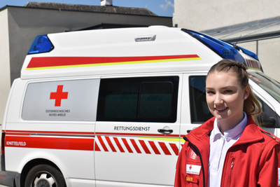 Sanitäterin Julia steht vor einem Rettungswagen und trägt die markante rote Uniformjacke.