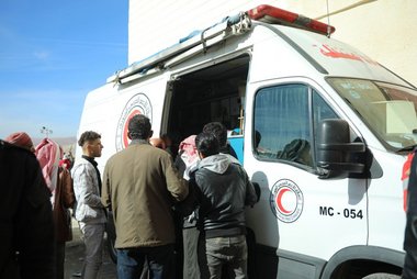 Eine Gruppe steht vor einem Auto des Syrisch-Arabischen Roten Halbmonds. Die Schiebetür seitlich ist geöffnet.