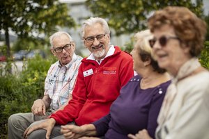 Ein Rotkreuz-Mitarbeiter sitzt mit seinen Klient:innen auf einer Parkbank und unterhält sich.