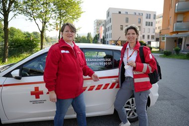 Michaela Schrumpf (Leiterin der Pflege und Betreuung, Rotes Kreuz Salzburg; links) und Marianne Zimmerebner (Leitung Mobile Krankenpflege, Rotes Kreuz Salzburg) stehen vor einem Fahrzeug der Mobilen Krankenpflege und freuen sich über den Einsatz der Notfall-Tracker.
