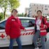Michaela Schrumpf (Leiterin der Pflege und Betreuung, Rotes Kreuz Salzburg; links) und Marianne Zimmerebner (Leitung Mobile Krankenpflege, Rotes Kreuz Salzburg) stehen vor einem Fahrzeug der Mobilen Krankenpflege und freuen sich über den Einsatz der Notfall-Tracker.