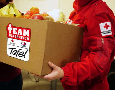 Rotkreuz-Mitarbeiterin trägt Schachtel mit Team Österreich Tafel Logo darauf. Schachtel ist voll mit Lebensmitteln.