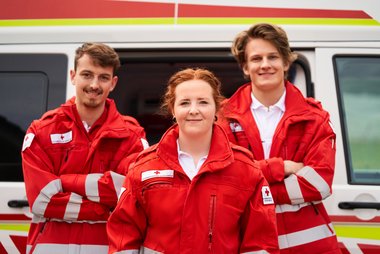 drei freiwillige Mitarbeiter:innen aus der Sparte Rettungsdienst vo einem Rettungsauto.