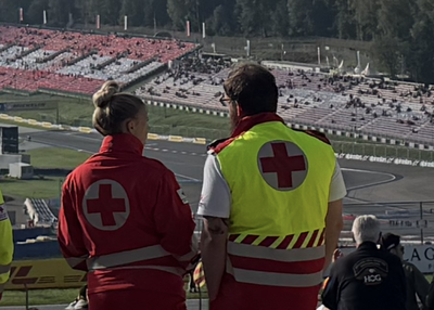 Zwei Rotkreuz-Mitarbeiter:innen stehen in Uniform am Red Bull Ring und überblicken das Geschehen auf den Tribünen und der Rennstrecke.