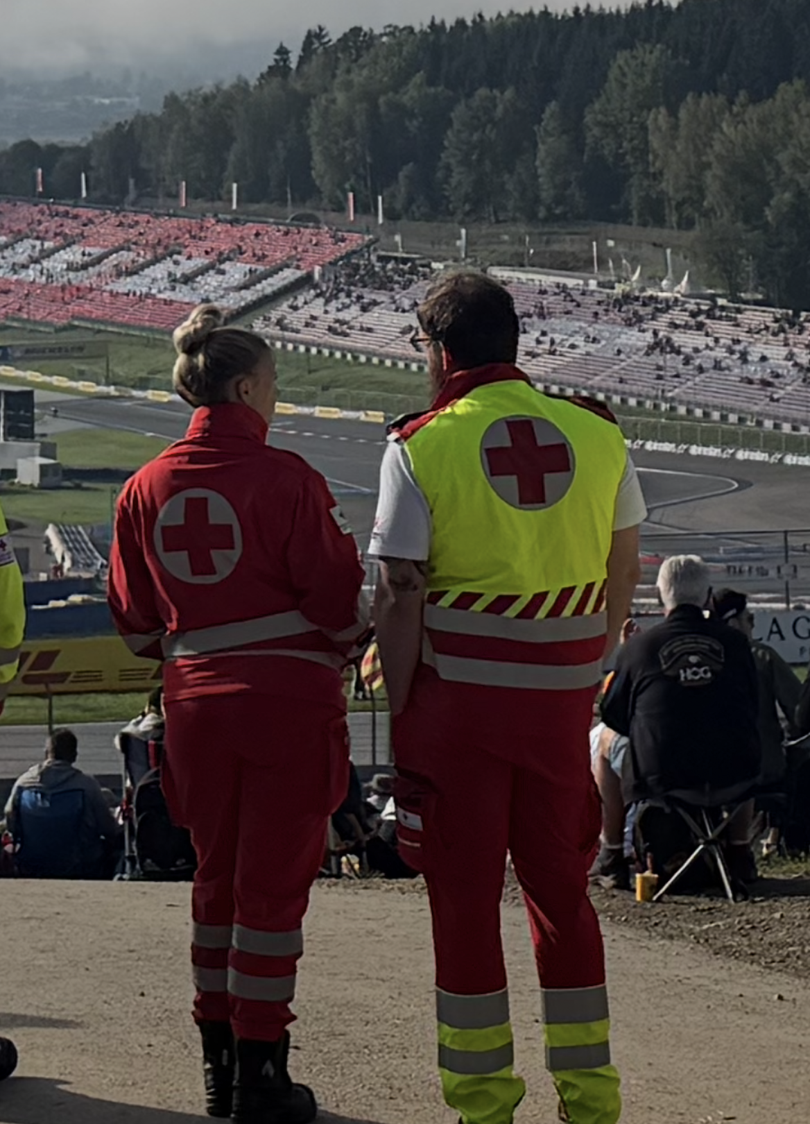 Zwei Rotkreuz-Mitarbeiter:innen stehen in Uniform am Red Bull Ring und überblicken das Geschehen auf den Tribünen und der Rennstrecke.