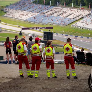 Man sieht Sanitäter:innen von hinten. Sie stehen erhöht und überblicken einen Teil des Red Bull Rings. Es sind auch einige Motorsportfans im Bild.