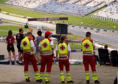 Man sieht Sanitäter:innen von hinten. Sie stehen erhöht und überblicken einen Teil des Red Bull Rings. Es sind auch einige Motorsportfans im Bild.