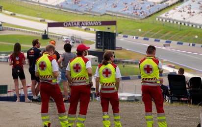 Man sieht Sanitäter:innen von hinten. Sie stehen erhöht und überblicken einen Teil des Red Bull Rings. Es sind auch einige Motorsportfans im Bild.