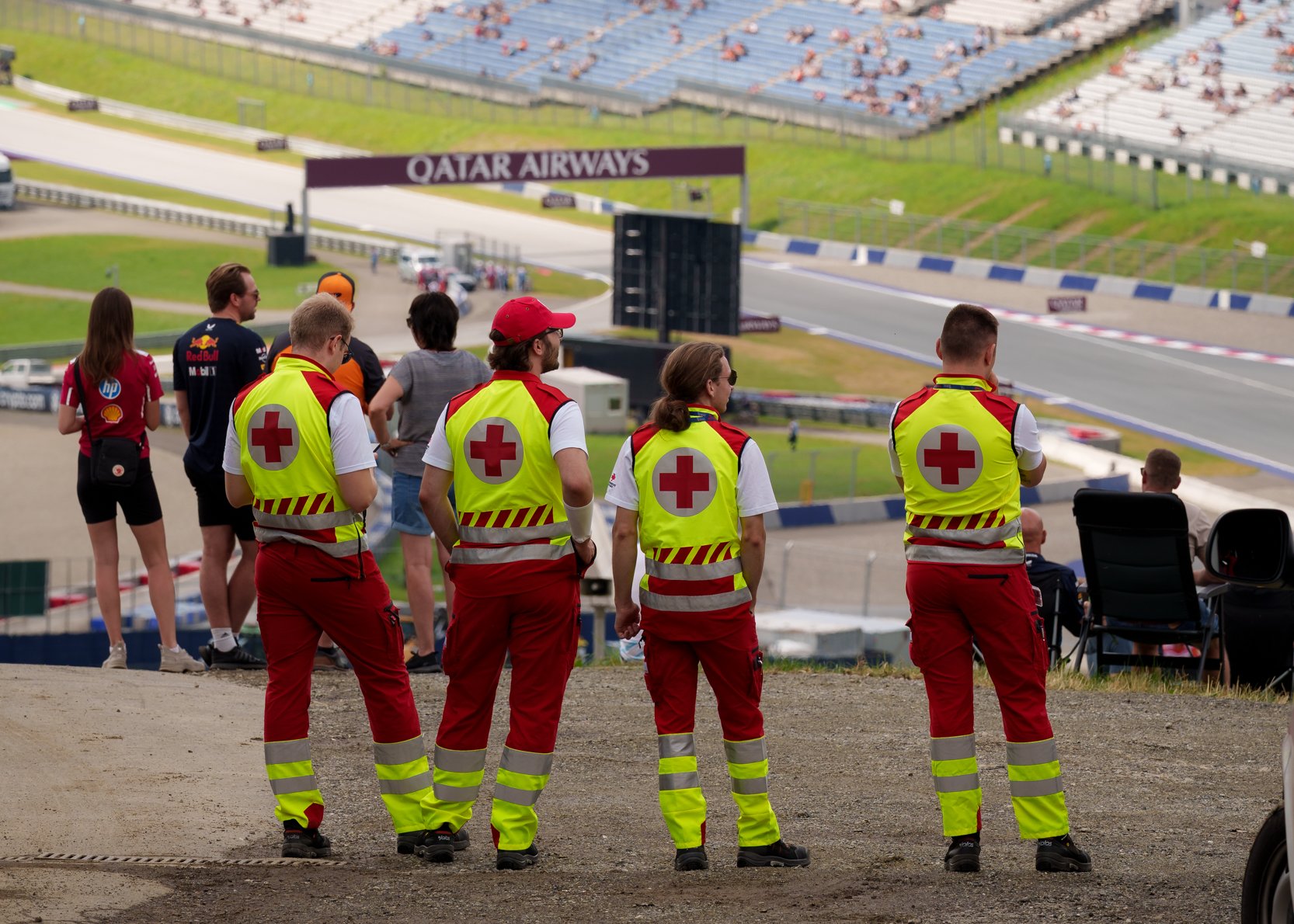 Man sieht Sanitäter:innen von hinten. Sie stehen erhöht und überblicken einen Teil des Red Bull Rings. Es sind auch einige Motorsportfans im Bild.