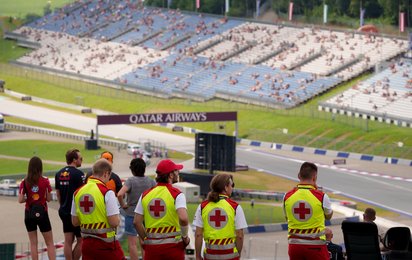 Man sieht Sanitäter:innen von hinten. Sie stehen erhöht und überblicken einen Teil des Red Bull Rings. Es sind auch einige Motorsportfans im Bild.