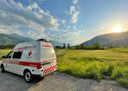 Ein Krankentransportwagen des Roten Kreuzes Waidhofen/Ybbs steht auf einen Schotterparkplatz wo im Hintergrund die Sonne hinter den Bergen untergeht.