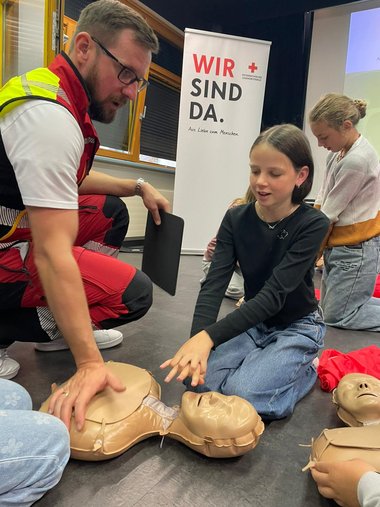 Ein Notarzt kniet gemeinsam mit einer Schülerin neben einer Übungspuppe und erklärt ihr die richtigen Handgriffe für die Herzmassage.