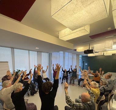 Wir sehen eine Gruppe Senioren auf Ihren Stühlen mit ausgestreckten Armen. Die Gruppe sitzt im Kreis und hat Freude an der Bewegung. Rotes Kreuz Salzburg: Seniorenwohnhaus Hallein 