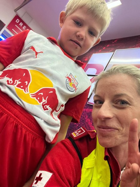 Aufnahme von unten. Links steht ein junger Bub in einem weiß-rotem FC-Red-Bull-Salzburg-Trikot, rechts eine Frau mit roter Rotkreuz-Jacke und gelber Weste darüber.