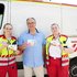Zwei Sanitäterinnen und ein Mann stehen vor einem Rettungswagen und halten Postkarten mit Hitze-Tipps in ihren Händen