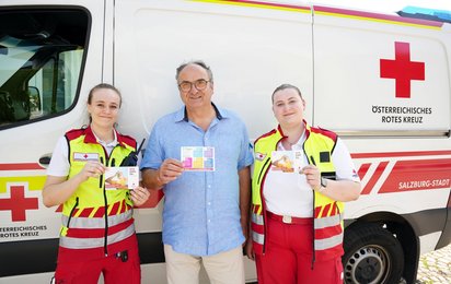 Zwei Sanitäterinnen und ein Mann stehen vor einem Rettungswagen und halten Postkarten mit Hitze-Tipps in ihren Händen