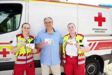 Zwei Sanitäterinnen und ein Mann stehen vor einem Rettungswagen und halten Postkarten mit Hitze-Tipps in ihren Händen