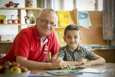 ein Freiwilliger unterstützt ein Kind beim Lesen, beide lächeln in die Kamera