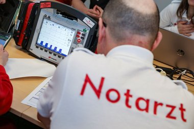 Blick auf den Bildschirm eines Corpuls-Gerätes, davor ein Notarzt von hinten in einem Polo-Shirt mit dem Aufdruck "Notarzt".