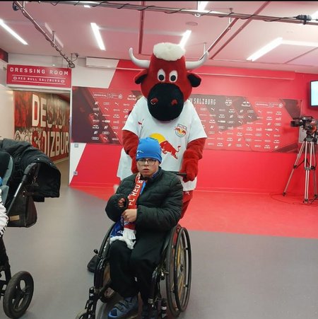 Das Maskottchen des FC Red Bull Salzburg steht hinter einem im Rollstuhl sitzenden Buben.