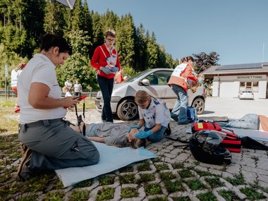 Eine Jugendliche übt die Versorgung einer verunfallten Person im Rahmen des Jugendrotkreuz-Landesbewerbs, im Hintergrund trainieren weitere Kinder und Jugendliche