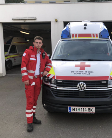 Kosovar Hoti steht in Rotkreuz-Uniform schräg vor einem Rettungswagen und lehnt sich leicht mit seinem linken Arm an der Motorhaube an.