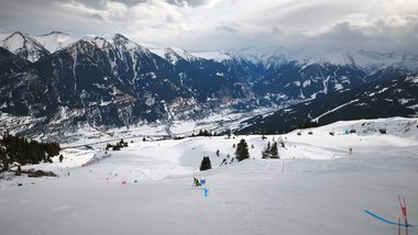 Panoramaaufnahme des Skigebiets in Bad Hofgastein. Im Vordergrund ist ein Rennkurs ausgesteckt, der von einem Skifahrer befahren wird. Im Hintergrund sind Berge und ein bewölkter Himmel zu sehen.