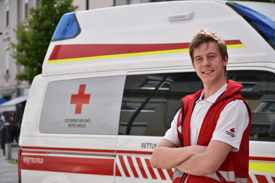Sanitäter Mario steht vor einem Rettungswagen und trägt die markante rote Uniformjacke.