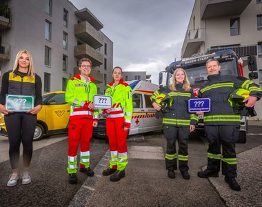 Einsatzkräfte der Feuerwehr und des Roten Kreuzes sowie eine Postmitarbeiterin stehen vor ihren Fahrzeugen und machen auf die Notwendigkeit aufmerksam, dass die Häuser gut beschildern sind