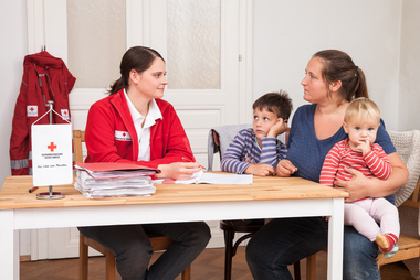 Eine Mutter mit zwei kleinen Kindern sitzt am Tisch mit einer Rotkreuz-Mitarbeiterin und lässt sich beraten.