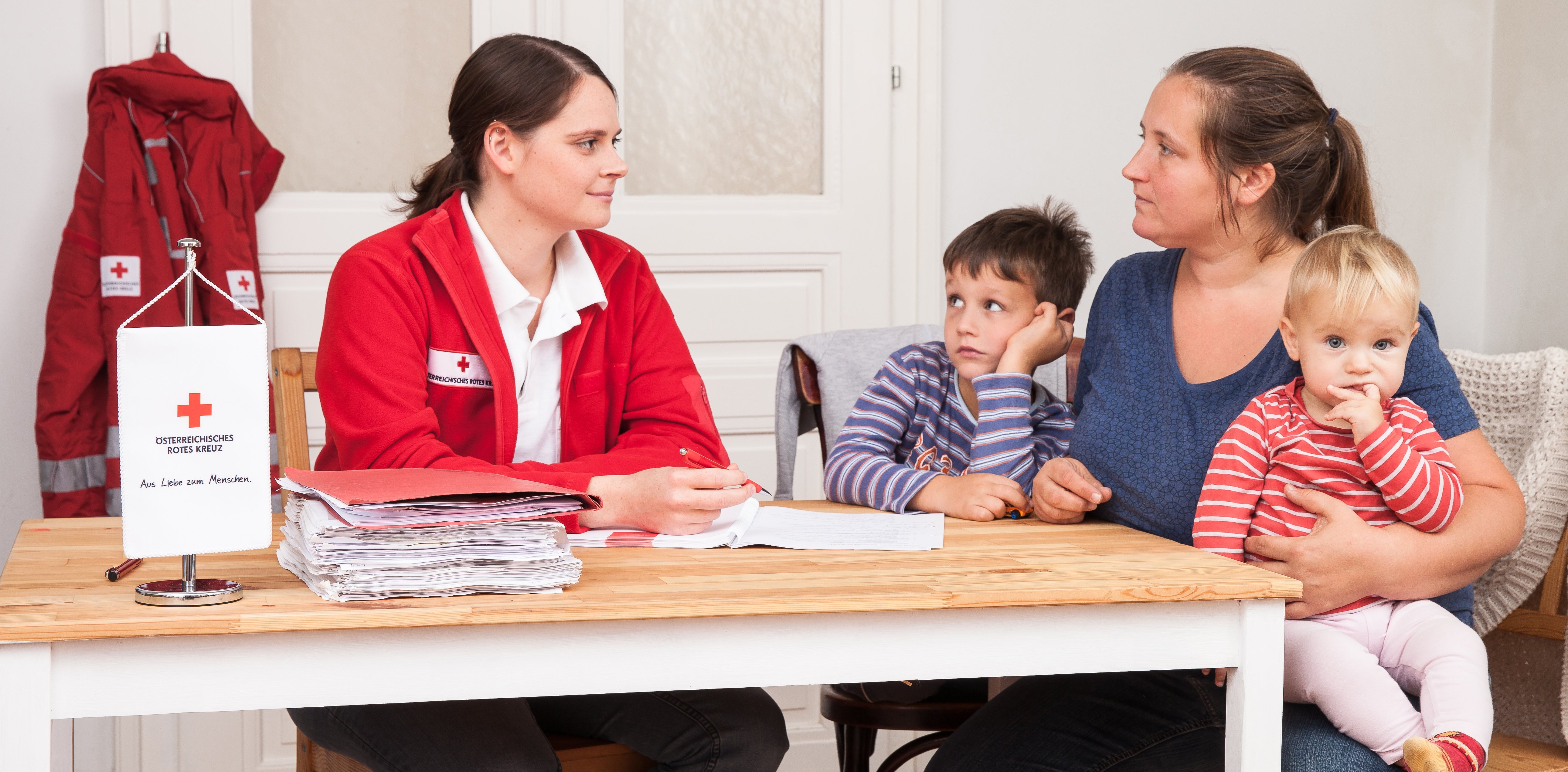 Eine Mutter mit zwei kleinen Kindern sitzt am Tisch mit einer Rotkreuz-Mitarbeiterin und lässt sich beraten.