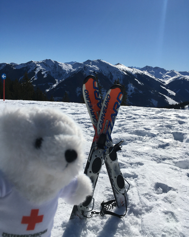 Bärli vor Skiern, die überkreuz im Schnee stecken. Er kennzeichnet so eine Unfallstelle auf der Skipiste.