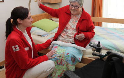 Eine ältere Dame sitzt aufrecht auf ihrem Pflegebett, eine Rotkreuz-Mitarbeiterin hockt vor ihr und unterstützt sie beim Aufstehen.
