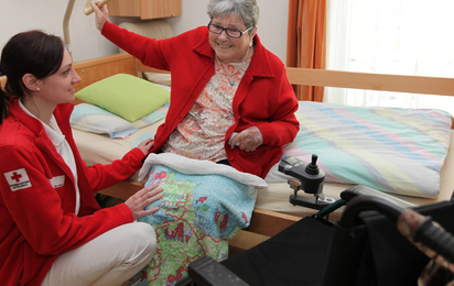 Eine ältere Dame sitzt aufrecht auf ihrem Pflegebett, eine Rotkreuz-Mitarbeiterin hockt vor ihr und unterstützt sie beim Aufstehen.