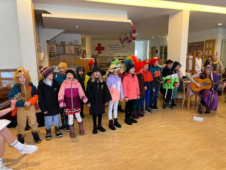 Gruppenfoto von verkleideten Kindern der Volksschule Sankt Gilgen, die Faschingslieder für die Bewohnerinnen singen. Daneben sitzt die Klassenlehrerin, die die Kinder mit ihrer Gitarre begleitet.