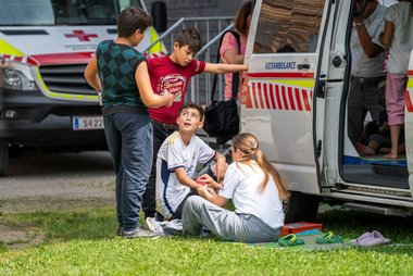 Eine Gruppe von Kindern sitzt beziehungsweise steht in einer Wiese vor der geöffneten Seitentür der Kidsambulanz