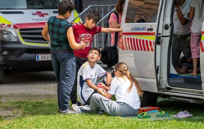 Eine Gruppe von Kindern sitzt beziehungsweise steht in einer Wiese vor der geöffneten Seitentür der Kidsambulanz