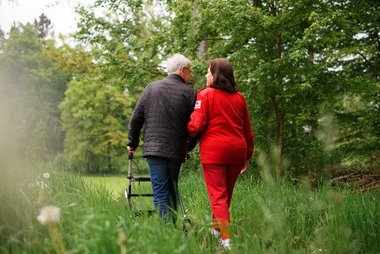 Ein älterer Mann mit Rollator spaziert in Begleitung einer Rotkreuz Mitarbeiterin auf einem Feldweg.
