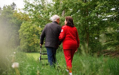 © Credit: OÖRK / Karin Lohberger Photography Ein älterer Mann mit Rollator spaziert in Begleitung einer Rotkreuz Mitarbeiterin auf einem Feldweg.